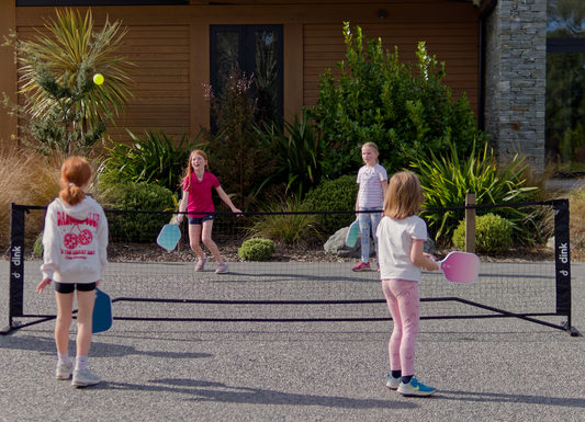 Children playing pickleball on an driveway with a house and plants in the background.